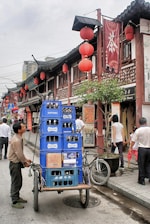 A bustling street scene featuring a man pushing a cart loaded with blue crates of Suntory bottles. The street is lined with traditional Chinese architecture, adorned with red lanterns and Chinese signage. Pedestrians are walking by, engaging in various activities. A bicycle is parked nearby, adding to the urban atmosphere.