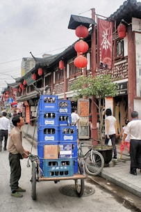 A bustling street scene featuring a man pushing a cart loaded with blue crates of Suntory bottles. The street is lined with traditional Chinese architecture, adorned with red lanterns and Chinese signage. Pedestrians are walking by, engaging in various activities. A bicycle is parked nearby, adding to the urban atmosphere.