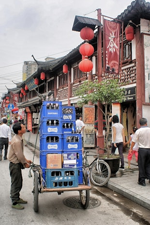 A bustling street scene featuring a man pushing a cart loaded with blue crates of Suntory bottles. The street is lined with traditional Chinese architecture, adorned with red lanterns and Chinese signage. Pedestrians are walking by, engaging in various activities. A bicycle is parked nearby, adding to the urban atmosphere.