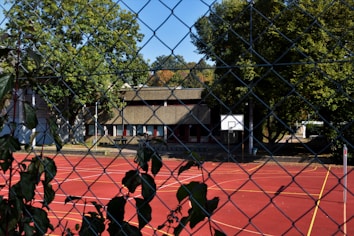 A red outdoor basketball court is visible through a chain-link fence. The court has a white basketball hoop and yellow lines marking the playing area. Surrounding the court are leafy green trees, and in the background, there is a building with a flat roof and several windows.