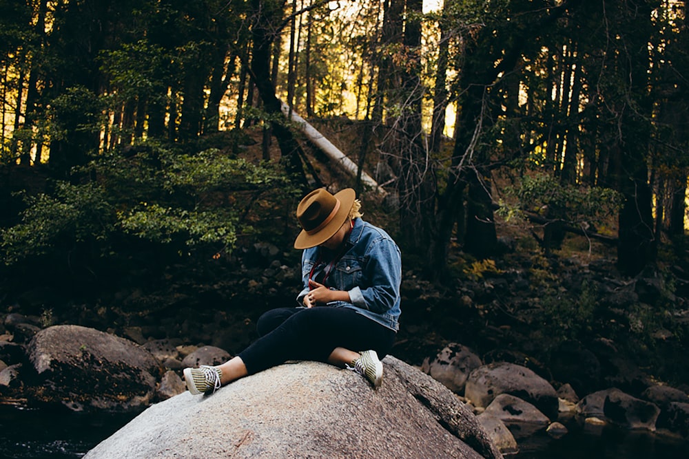 person wearing brown hat sitting on gray rock near trees during daytime