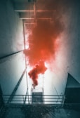 A narrow, tall space with white walls and a metal railing, where thick plumes of red smoke fill the center. The smoke contrasts sharply with the stark, cold ambiance created by the aged and slightly peeling walls.