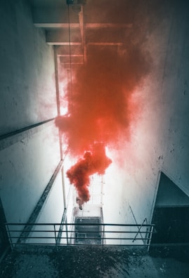 A narrow, tall space with white walls and a metal railing, where thick plumes of red smoke fill the center. The smoke contrasts sharply with the stark, cold ambiance created by the aged and slightly peeling walls.