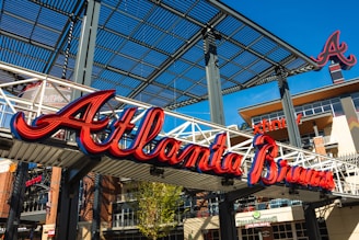 Large red and blue script signage reads 'Atlanta Braves' on a white metal structure. Surrounding area includes a modern architectural setting with brick and metal elements. Visible is an 'A' logo and parts of the structure, including horizontal beams and vertical girders.