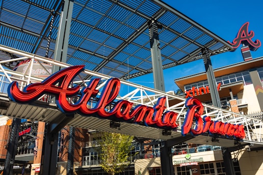 Large red and blue script signage reads 'Atlanta Braves' on a white metal structure. Surrounding area includes a modern architectural setting with brick and metal elements. Visible is an 'A' logo and parts of the structure, including horizontal beams and vertical girders.