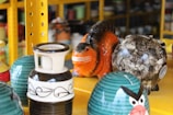 A collection of colorful ceramic objects arranged on yellow metal shelves. An orange creature with a wide grin sits among patterned vases and a round object decorated with abstract designs. A piece resembling a character with angry eyebrows features prominently in the foreground.