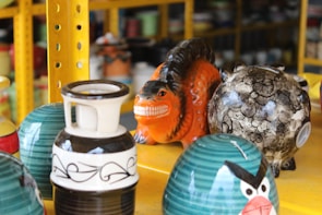A collection of colorful ceramic objects arranged on yellow metal shelves. An orange creature with a wide grin sits among patterned vases and a round object decorated with abstract designs. A piece resembling a character with angry eyebrows features prominently in the foreground.