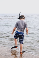 A young person is wearing snorkeling gear and standing at the edge of a body of water, about to step in. They are equipped with a snorkel mask, fins, and what appears to be a wetsuit or swimming attire. The surroundings suggest a calm, shallow coastal area, with gentle waves lapping at the rocky shore.