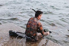 A person wearing a camouflage wetsuit and snorkel gear sits by the edge of the water. The individual appears to be preparing for diving or spearfishing, with fins and a speargun at their side.