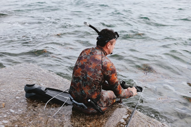 A person wearing a camouflage wetsuit and snorkel gear sits by the edge of the water. The individual appears to be preparing for diving or spearfishing, with fins and a speargun at their side.