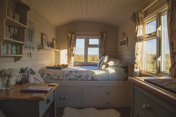 An empty bed sits beside a set of windows in a small, sunlit bedroom during the daytime.