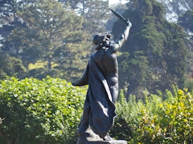 A bronze statue of a warrior figure holding a sword aloft, standing amidst lush green foliage. The figure is draped in a cloak and set against a backdrop of dense, towering trees, casting shadows that contrast with the bright sunlight illuminating the scene.