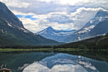A serene mountain landscape featuring a tranquil lake reflecting the surrounding snow-capped peaks. The sky is partly cloudy with patches of blue breaking through, adding depth and drama to the scene. Dense forests border the lake, enhancing the natural beauty of the environment.