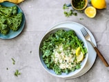 A bowl filled with white rice topped with fresh, chopped greens and broccolini florets, accompanied by lemon wedges. A fork rests on the side of the plate, which is placed on a gray stone textured surface. Nearby, there’s a plate with more broccolini, a cup of chopped herbs, and half a lemon.
