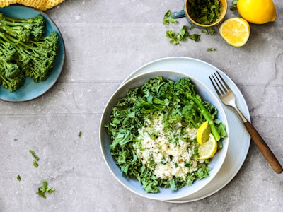 A bowl filled with white rice topped with fresh, chopped greens and broccolini florets, accompanied by lemon wedges. A fork rests on the side of the plate, which is placed on a gray stone textured surface. Nearby, there’s a plate with more broccolini, a cup of chopped herbs, and half a lemon.