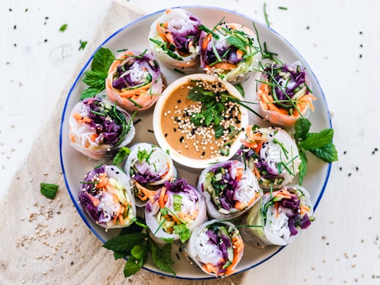 A platter of fresh Vietnamese spring rolls arranged in a circular pattern around a small bowl of dipping sauce. The rolls are filled with an assortment of colorful vegetables, including purple cabbage, carrots, and green herbs. The sauce is garnished with sesame seeds and fresh mint leaves. There are sprigs of mint and scattered sesame seeds on the white surface around the plate.