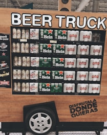A display resembling a beer truck features various brands of beer, including Sol, Heineken, and Schneider. The structure mimics a vehicle with a wheel at the bottom and wooden paneling. It includes a promotional slogan written in Spanish.