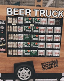 A display resembling a beer truck features various brands of beer, including Sol, Heineken, and Schneider. The structure mimics a vehicle with a wheel at the bottom and wooden paneling. It includes a promotional slogan written in Spanish.