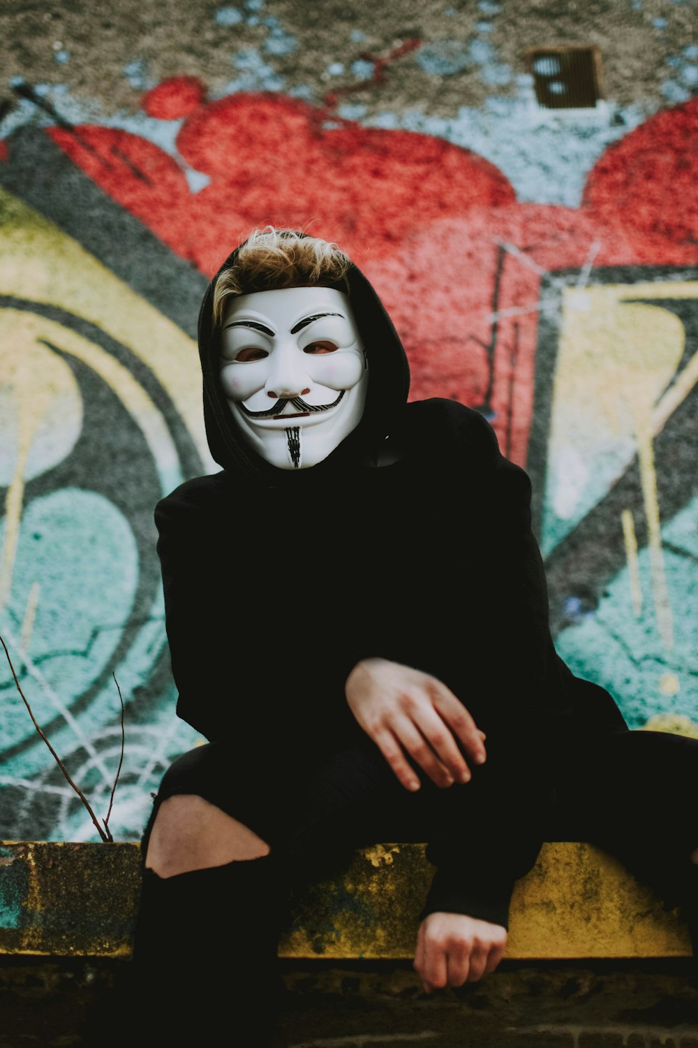 man wearing Guy-Fawkes mask sitting on yellow concrete bench