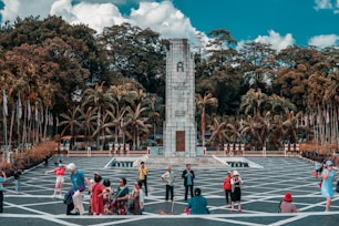A tall stone monument stands in the center of an open plaza, surrounded by lush greenery and tall palm trees. The area is filled with people, many of whom are tourists walking around, taking photos, and interacting with each other. Numerous flags line the side of the plaza, adding a sense of national pride and ceremony. The sky above is bright with white clouds contrasting against a blue sky.