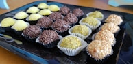 An assortment of spherical sweets placed on a black tray. The candies are arranged in rows and each row comprises a different type of confectionery with varying textures and coatings: the first row has sweets covered in yellow granulated sugar, the second has chocolate-covered sweets, the third row consists of sweets with a green powder-like coating, and the last row includes sweets covered in chopped nuts.