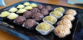 An assortment of spherical sweets placed on a black tray. The candies are arranged in rows and each row comprises a different type of confectionery with varying textures and coatings: the first row has sweets covered in yellow granulated sugar, the second has chocolate-covered sweets, the third row consists of sweets with a green powder-like coating, and the last row includes sweets covered in chopped nuts.