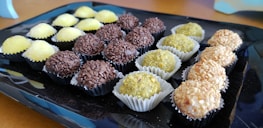 An assortment of spherical sweets placed on a black tray. The candies are arranged in rows and each row comprises a different type of confectionery with varying textures and coatings: the first row has sweets covered in yellow granulated sugar, the second has chocolate-covered sweets, the third row consists of sweets with a green powder-like coating, and the last row includes sweets covered in chopped nuts.