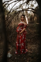 A pregnant woman stands among arching tree branches in a forest, wearing a long red floral dress. Dappled sunlight filters through the trees, highlighting her figure and casting soft shadows on the ground. The atmosphere is serene and natural, with an earthy, rustic setting.
