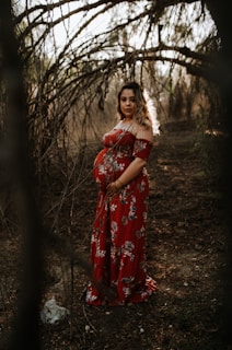 A pregnant woman stands among arching tree branches in a forest, wearing a long red floral dress. Dappled sunlight filters through the trees, highlighting her figure and casting soft shadows on the ground. The atmosphere is serene and natural, with an earthy, rustic setting.