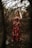 A pregnant woman stands among arching tree branches in a forest, wearing a long red floral dress. Dappled sunlight filters through the trees, highlighting her figure and casting soft shadows on the ground. The atmosphere is serene and natural, with an earthy, rustic setting.