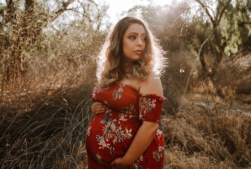 A pregnant woman stands in a sunlit field, surrounded by tall dry grass and trees. She wears a red floral dress and holds her belly. The natural lighting gives her hair a glowing effect.