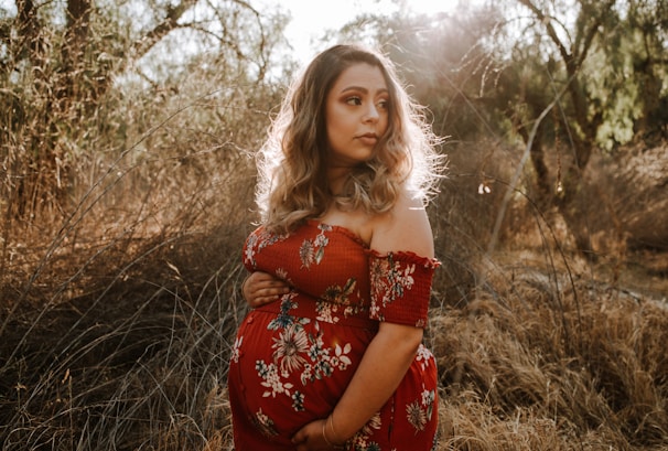 A pregnant woman stands in a sunlit field, surrounded by tall dry grass and trees. She wears a red floral dress and holds her belly. The natural lighting gives her hair a glowing effect.
