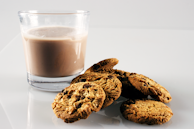 A glass of chocolate milk is placed on a smooth white surface next to a small pile of chocolate chip cookies. The cookies have a golden-brown color with visible chocolate chips.