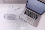 A laptop is placed on a white desk displaying a logo for 'Sparkling Marketing'. Beside the laptop, a glass jar lies on its side with its contents—numerous small, clear beads—spilled across the surface. Nearby, a pen with a decorative gem sits on some business cards, contributing to a coordinated and stylish workspace aesthetic.