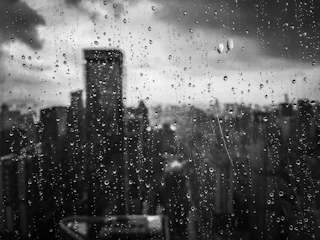 A black-and-white photograph of a mid-century noir cityscape at dusk, viewed through rain-speckled glass.