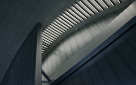 A modern architectural structure with a series of parallel fluorescent lights on the ceiling, casting a geometric pattern of light and shadow on the walls below. The design features clean lines and smooth, metallic surfaces, creating a sleek and futuristic appearance.