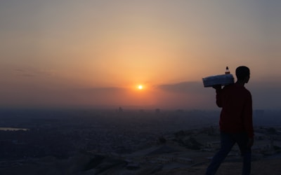 A silhouette of a person carrying a tray with bottles set against the backdrop of a vibrant sunset over a sprawling city landscape. The sky is painted with hues of orange, pink, and purple, while the sun casts a warm glow.