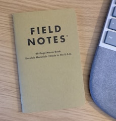 A Field Notes memo book with a brown cover lies on a wooden desk next to part of a computer keyboard. The memo book is labeled as a 48-page book made from durable materials in the USA.