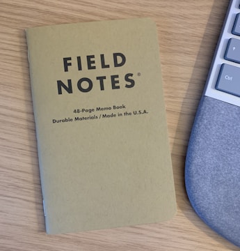A Field Notes memo book with a brown cover lies on a wooden desk next to part of a computer keyboard. The memo book is labeled as a 48-page book made from durable materials in the USA.