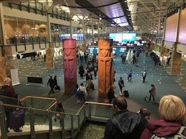 A busy airport terminal features large, carved wooden figures as central pieces of art. Travelers are walking in various directions, some with luggage. Information displays and check-in counters are visible in the background, with the ceiling showing an industrial architectural design.