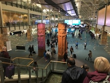A busy airport terminal features large, carved wooden figures as central pieces of art. Travelers are walking in various directions, some with luggage. Information displays and check-in counters are visible in the background, with the ceiling showing an industrial architectural design.