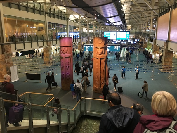 A busy airport terminal features large, carved wooden figures as central pieces of art. Travelers are walking in various directions, some with luggage. Information displays and check-in counters are visible in the background, with the ceiling showing an industrial architectural design.