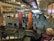 A busy airport terminal features large, carved wooden figures as central pieces of art. Travelers are walking in various directions, some with luggage. Information displays and check-in counters are visible in the background, with the ceiling showing an industrial architectural design.