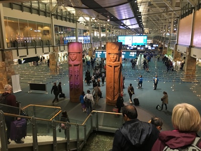 A busy airport terminal features large, carved wooden figures as central pieces of art. Travelers are walking in various directions, some with luggage. Information displays and check-in counters are visible in the background, with the ceiling showing an industrial architectural design.
