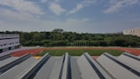 A large sports field with green grass is surrounded by a red athletics track. In the foreground, several modern buildings with pitched roofs are visible. A dense line of trees stretches in the middle ground, providing a lush green barrier between the sports field and the distant high-rise buildings. The sky is partly cloudy with patches of blue visible.
