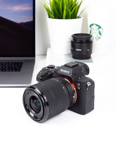 A Sony camera is positioned on a white surface with a laptop to its left, a Starbucks cup and plant in a white pot to its right. A Canon lens is also visible nearby, creating a minimalist and professional setup.