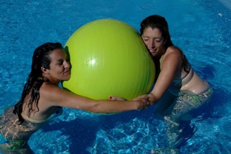Two women are in a swimming pool, each holding onto a large yellow exercise ball. Both women have wet hair and are wearing swimsuits, enjoying the water together.