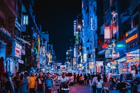 A bustling urban street at night, filled with people walking and vehicles passing by. Numerous bright neon signs in various languages illuminate the buildings, contributing to a vibrant nightlife atmosphere. Shops and restaurants line the street, with a diverse crowd engaging in leisure activities.
