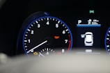 A car dashboard with a tachometer displaying RPM levels and warning lights illuminated, including oil and engine warnings. A digital screen shows the car in a parking mode with an open door and details like fuel range and comfort mode.