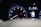 A car dashboard with a tachometer displaying RPM levels and warning lights illuminated, including oil and engine warnings. A digital screen shows the car in a parking mode with an open door and details like fuel range and comfort mode.